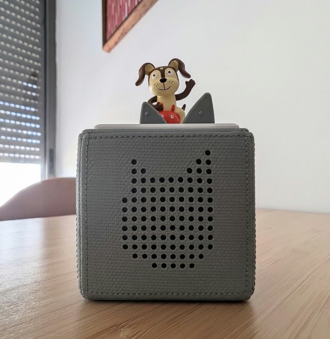 A grey Toniebox on a wooden table with a dog Tonie figurine standing on top of it